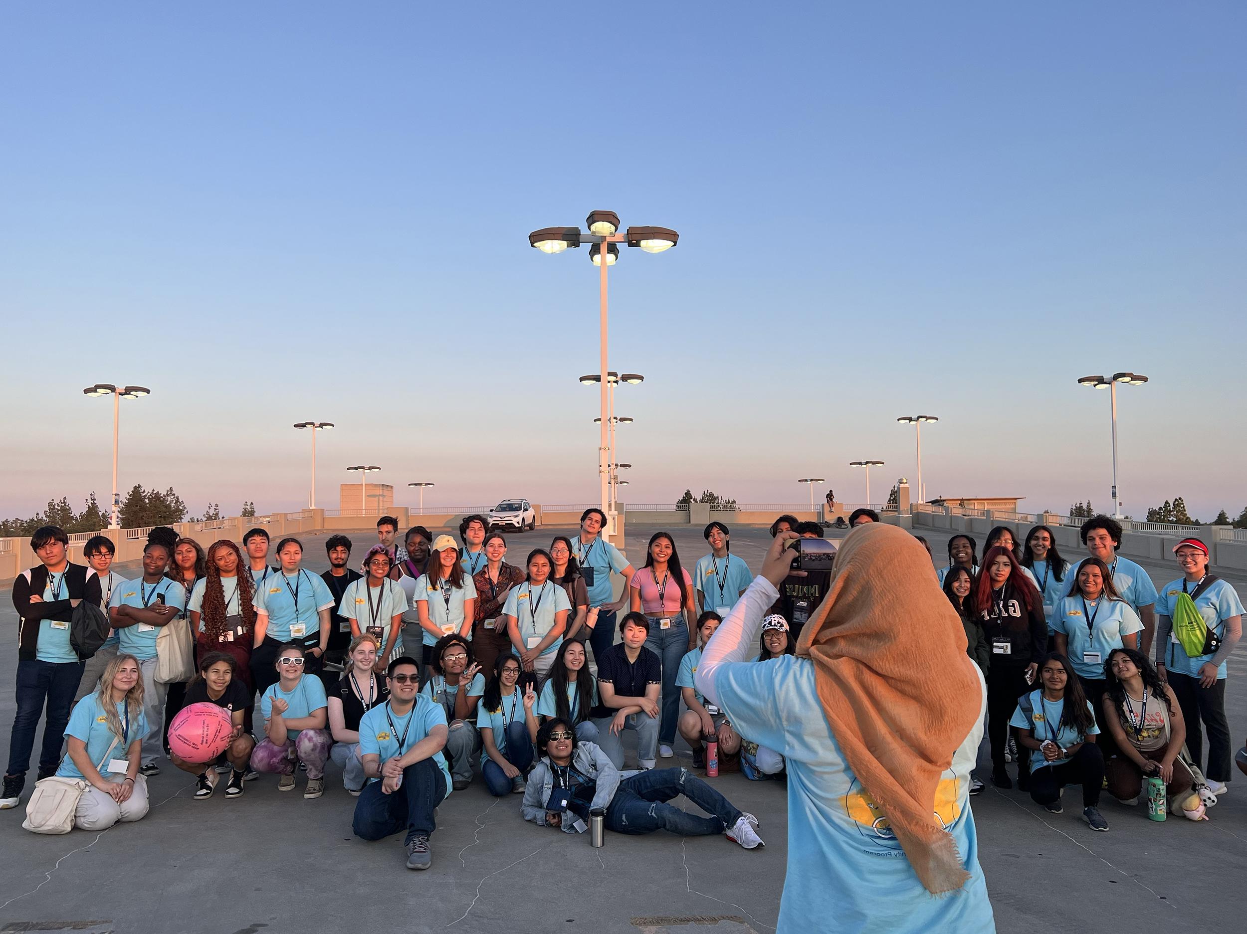 A woman wearing a hijab takes a group photo of students around sunset on campus